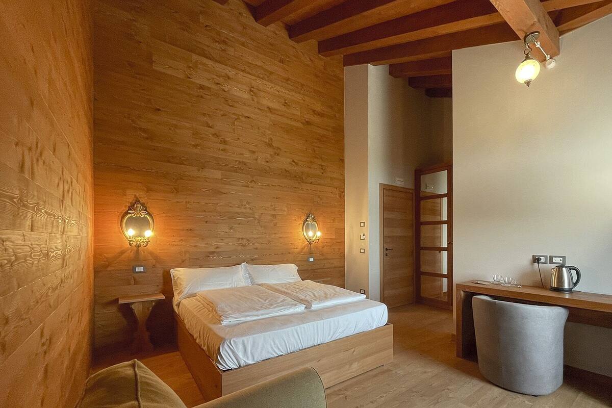 Una habitación acogedora con techo de madera, cama con sábanas blancas, dos apliques de pared, una mesita con un hervidor y un cubo de basura.