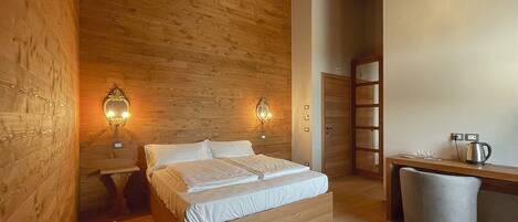 Una habitación acogedora con techo de madera, cama con sábanas blancas, dos apliques de pared, una mesita con un hervidor y un cubo de basura.