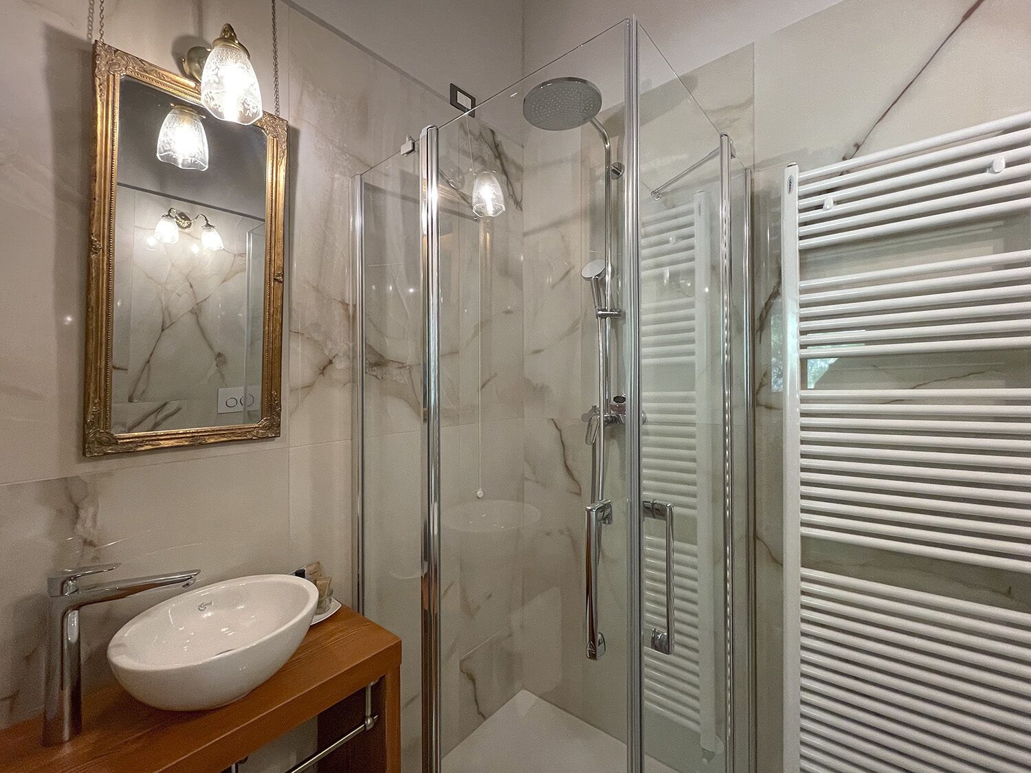 Een moderne badkamer met een douchecabine van glas, een wand betegeld met marmer, een houten wastafelmeubel met een witte wasbak en een grote spiegel met gouden lijst.