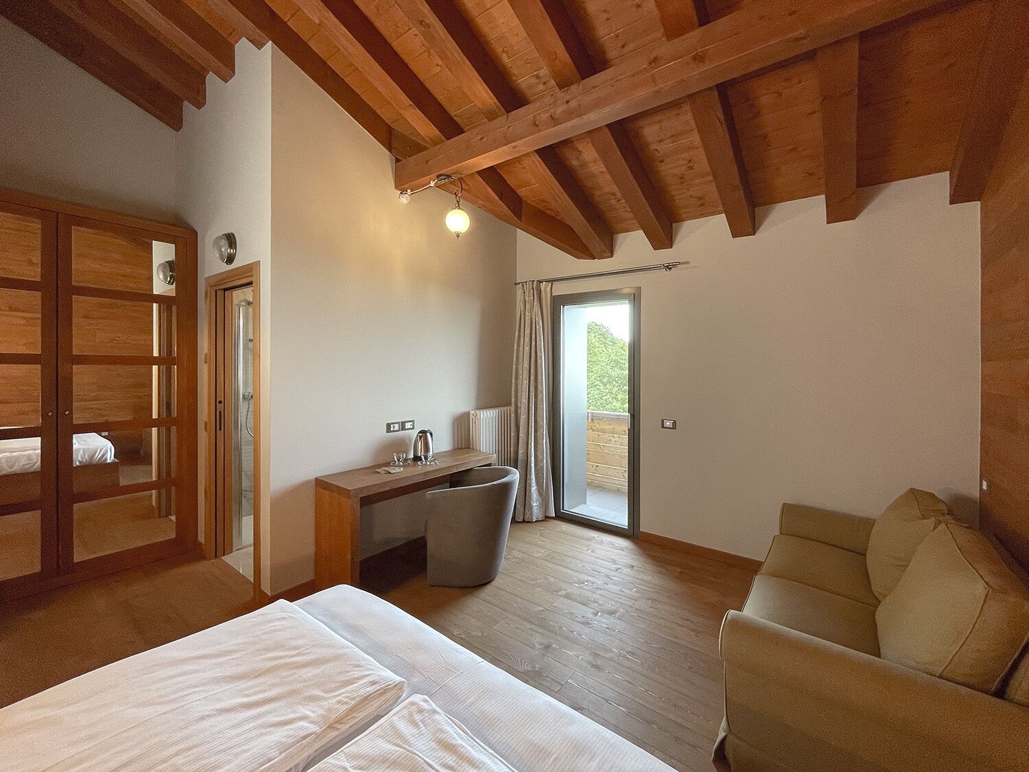 Ein Schlafzimmer mit Holzbalkendecke, einem Bett, einem kleinen Schreibtisch mit Stuhl und einem Fenster mit Blick ins Grüne.