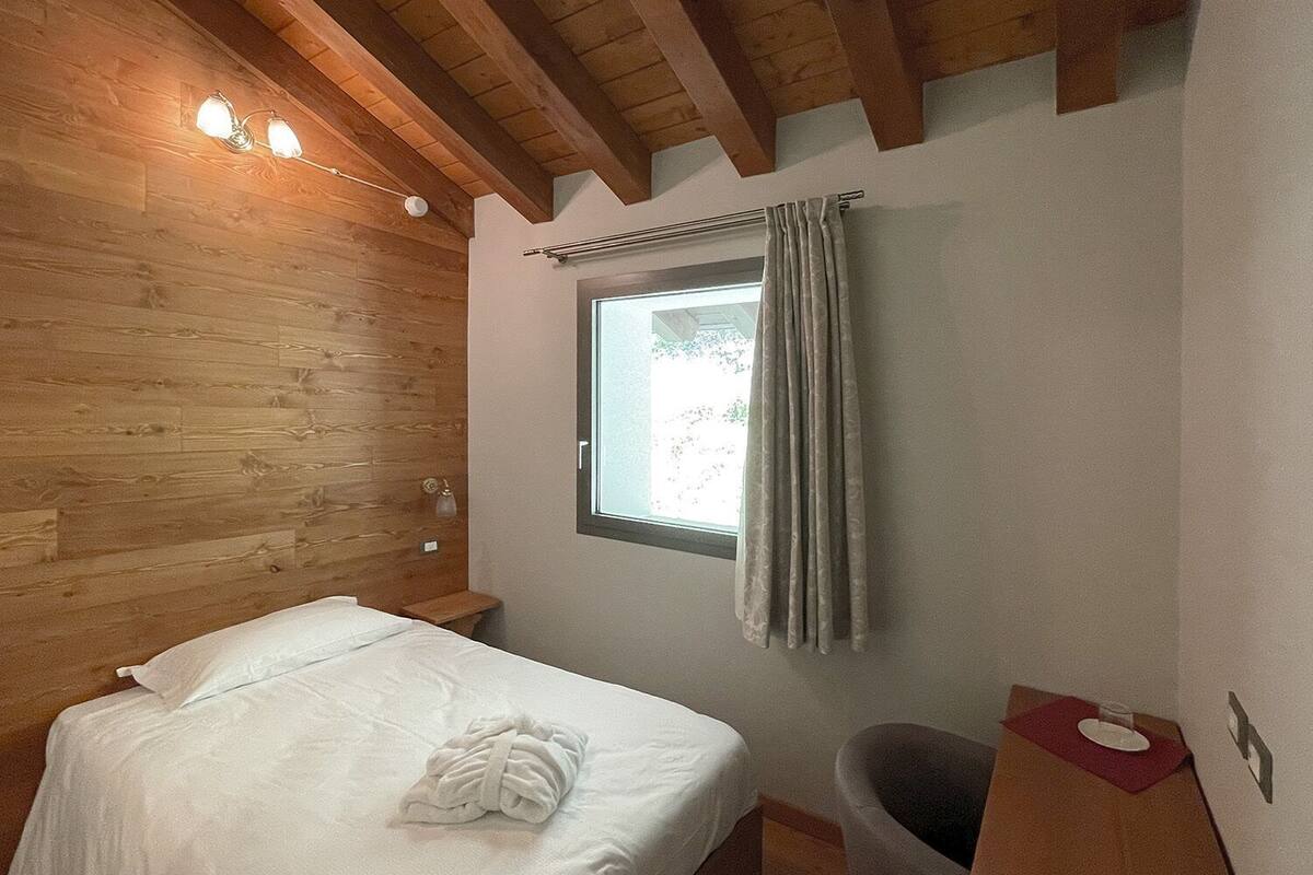 Un dormitorio con techo de madera, una cama con ropa de cama blanca, una ventana con cortinas, una mesita de noche y un cubo de basura.