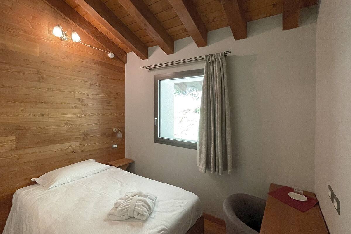 Un dormitorio con techo de madera, una cama individual con cubrecama blanco, una ventana con cortinas y una mesita de noche pequeña.
