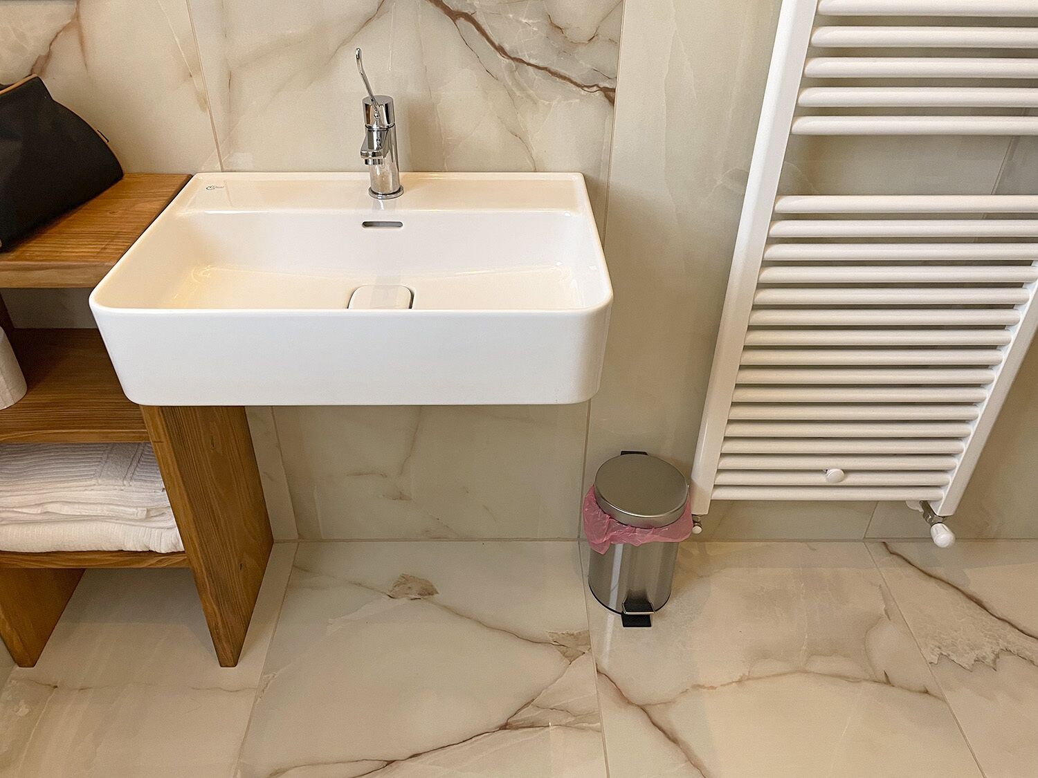 A bathroom with a white sink, a wooden shelf, a trash bin, and a white towel rack.