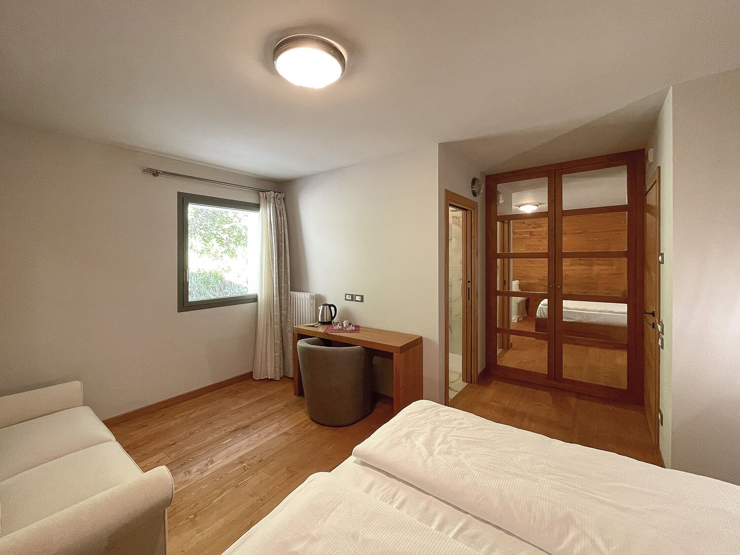 A hotel room with a bed, a desk with a chair, a window with curtains, a radiator, and a wooden door.
