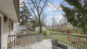A wooden deck with a wicker chair, overlooking a grassy yard with red chairs and a white building in the background.