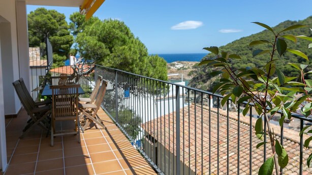 Un balcón con suelo de baldosas, una mesa y sillas, con vistas a un paisaje costero con árboles y el mar.