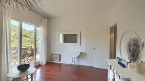 Amplia habitación con suelo de madera, una mesa de comedor con un cuenco, un radiador, un armario blanco y un amplio ventanal con vistas a zonas verdes.