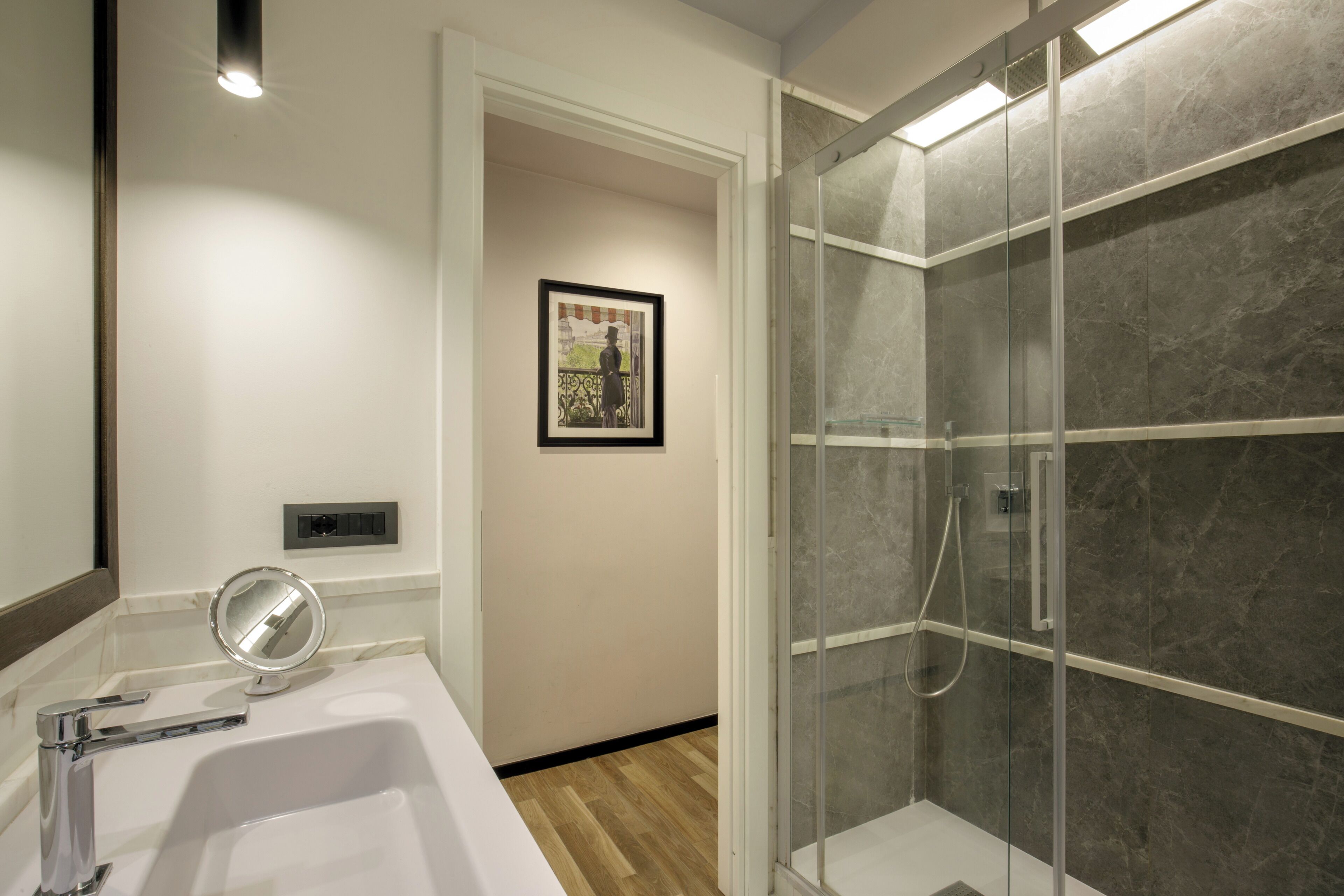 A bathroom with a white sink, a mirror, a framed picture on the wall, and a glass-enclosed shower area.