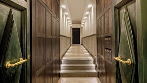 Un long couloir étroit, avec des portes en bois et un sol en marbre, qui mène à une porte sombre au bout.