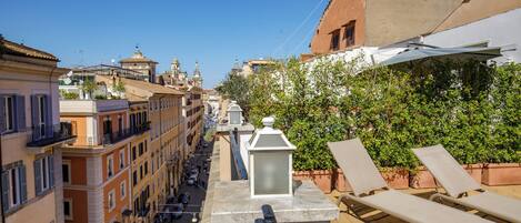 A rooftop terrace with lounge chairs, a potted plant, and a view of a city street.