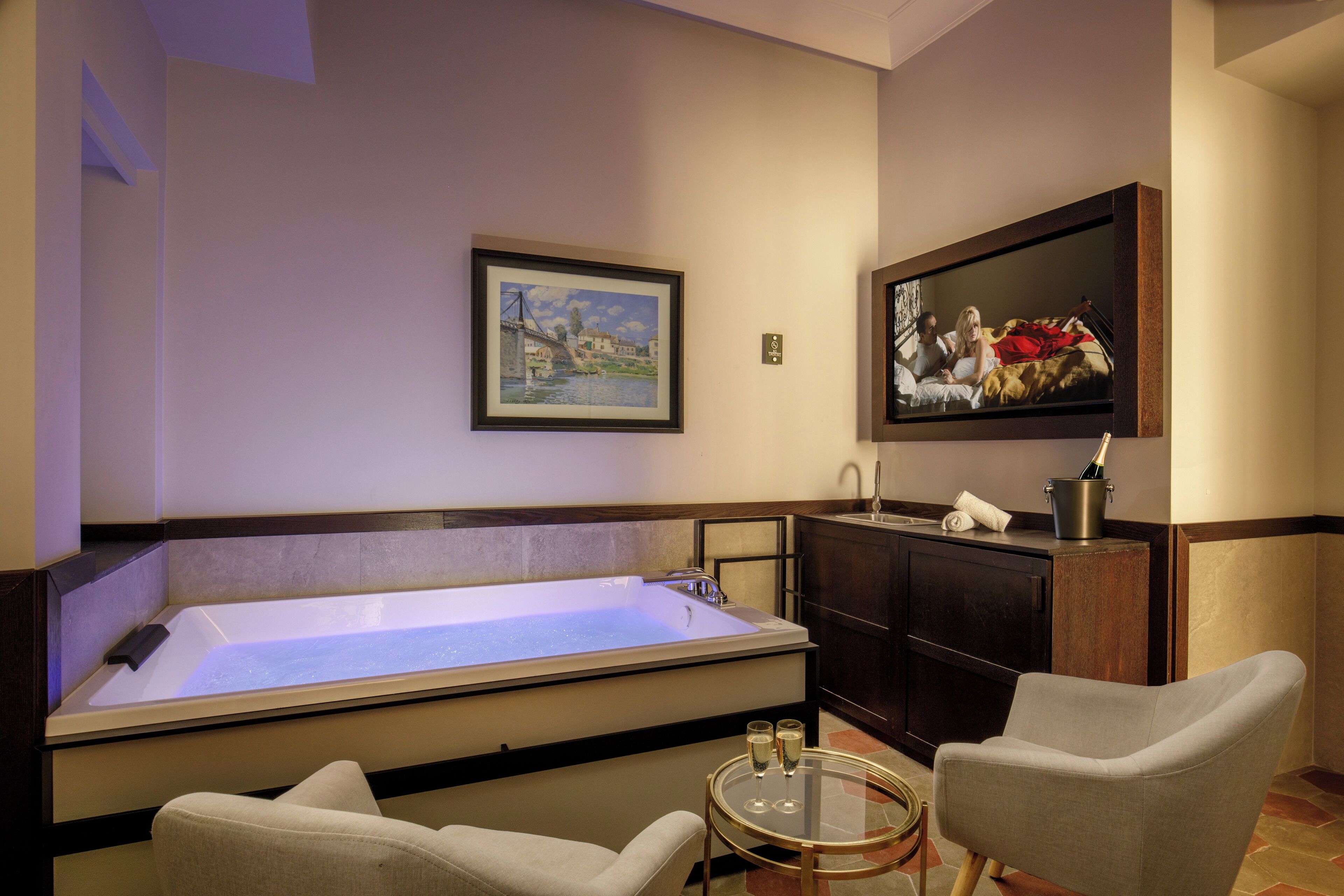 A room with a large bathtub, a small round table with glasses, two chairs, a cabinet, and two framed pictures on the wall.