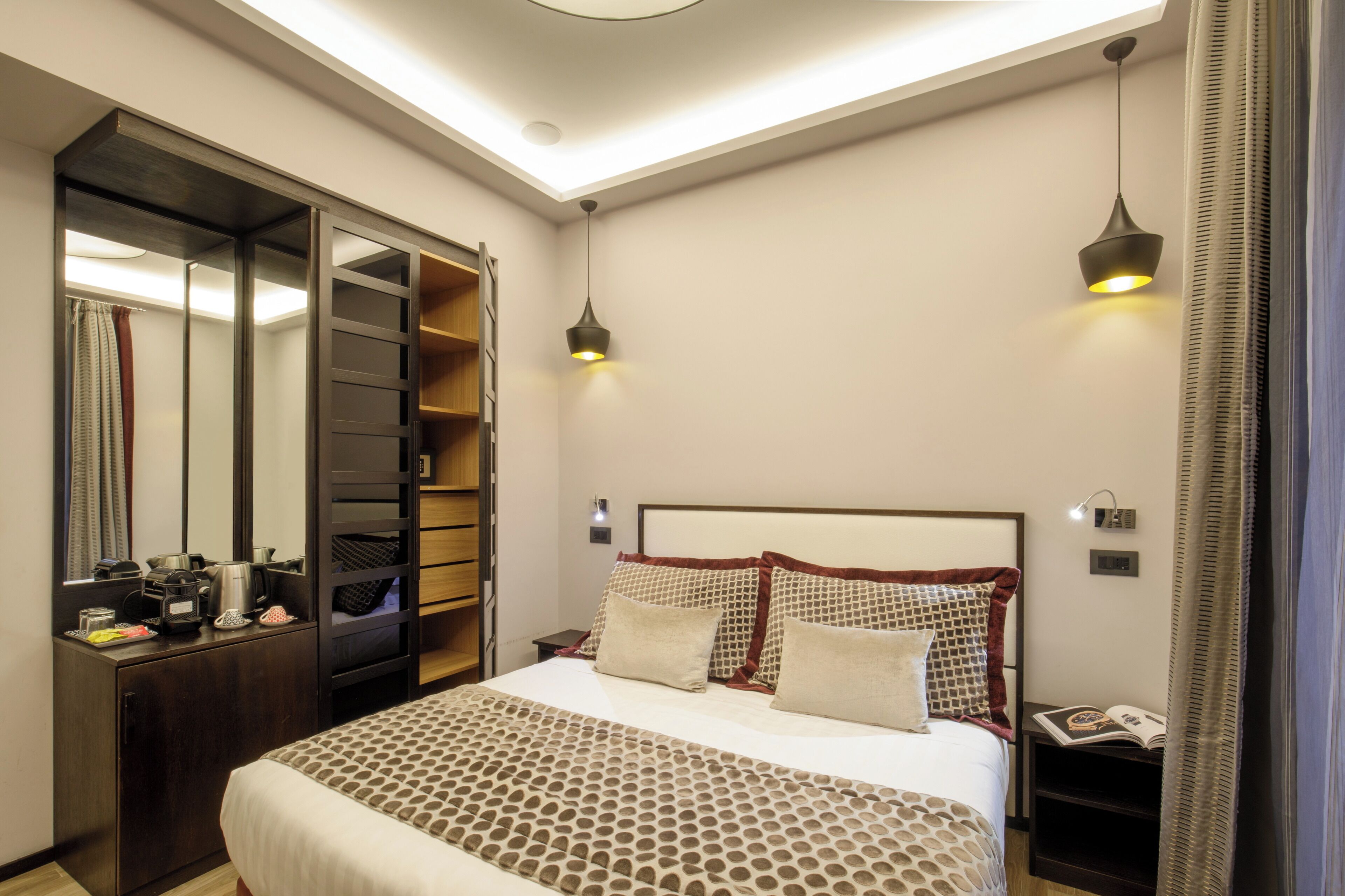 A neatly arranged hotel room with a large bed, a wardrobe with multiple shelves, a bedside table with a lamp, and a nightstand with a book.