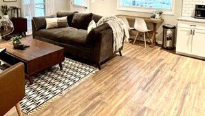 A cozy living room with a dark sofa, a coffee table, and a patterned rug. There is a dining area with a small table and chairs, and a kitchen area with cabinets and a trash can. - Dreamy Cottage Getaway w/ Sunset Lakeview (Cadiz)