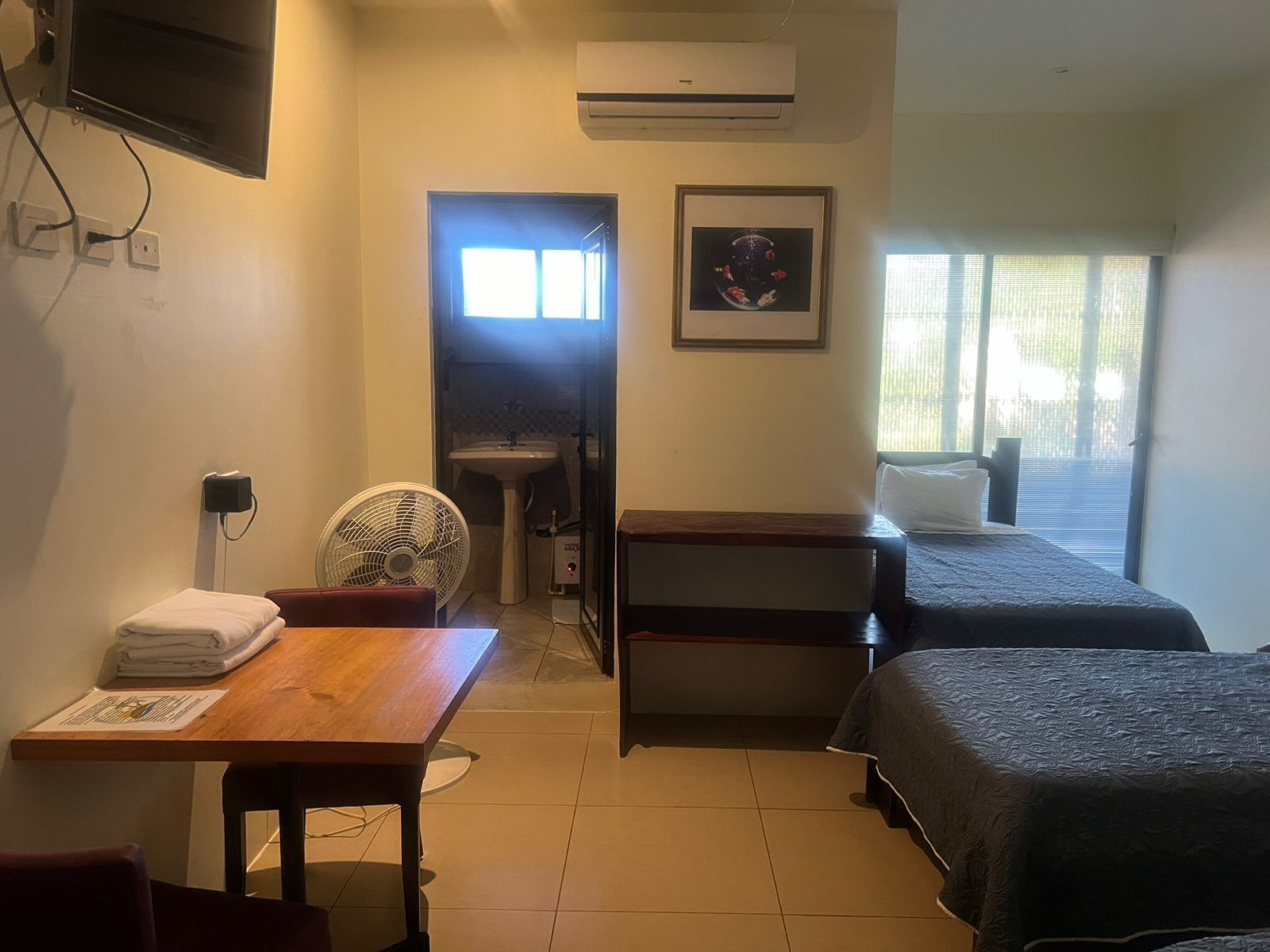 Habitación de hotel con un escritorio de madera, una silla, dos camas, un lavamanos y una ventana con persianas.