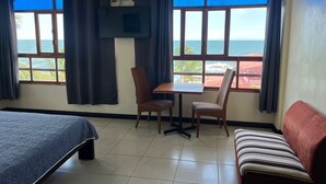 Quarto com uma cama, uma mesa com duas cadeiras, uma TV fixada na parede e grandes janelas com vista para o oceano.