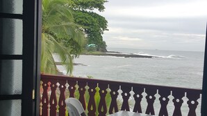 Varanda com mesa e cadeiras, vista para a costa com palmeiras e o oceano.