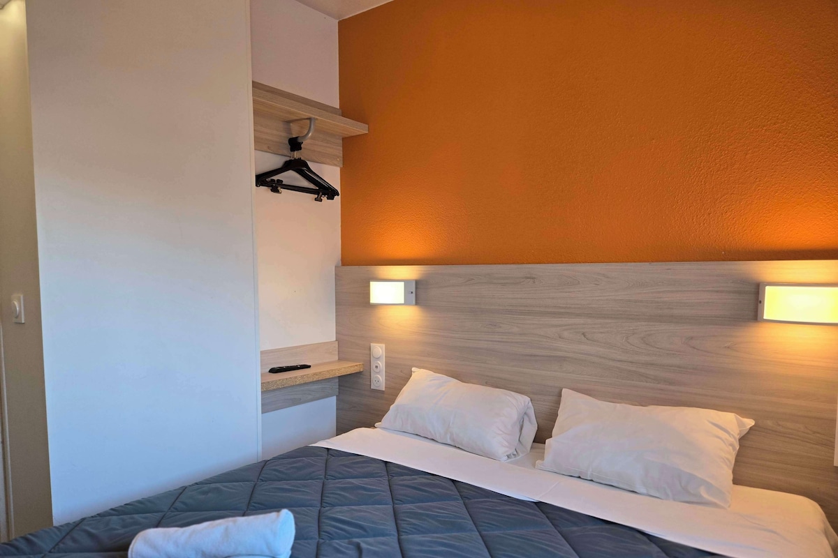 A neatly made bed with a grey quilt, white pillows, and a rolled towel on the left side. There is a wooden headboard with two wall-mounted lights on either side.