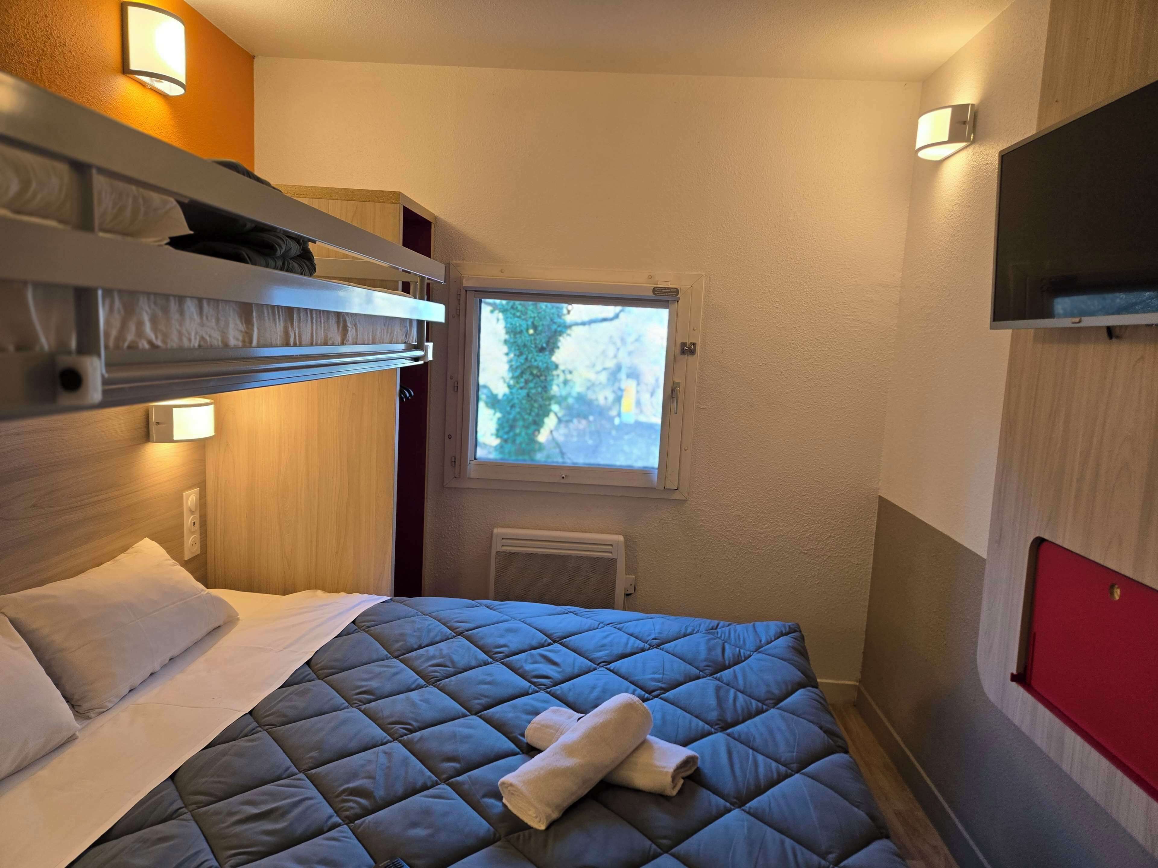 Une chambre d’hôtel compacte avec un lit superposé, une petite fenêtre, un téléviseur à écran plat et un radiateur.