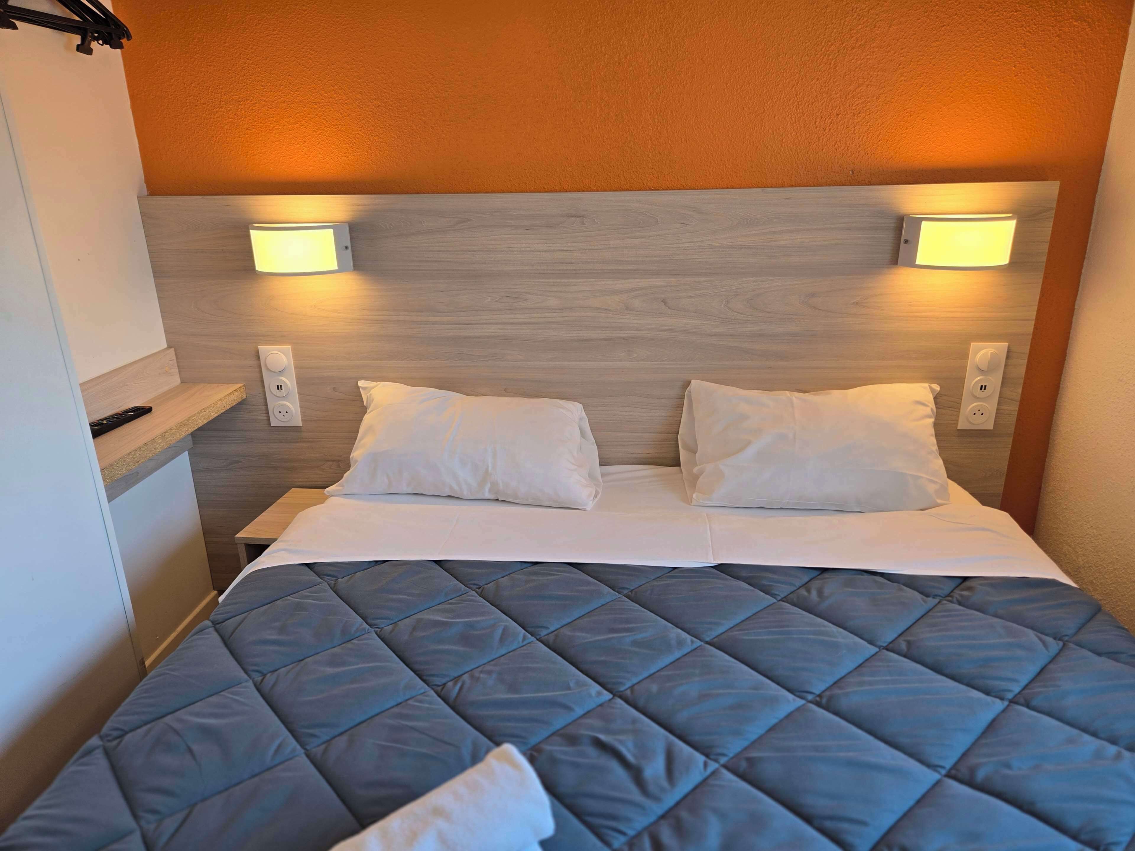 A hotel room with a bed, two bedside lamps, a nightstand, and a remote control on the nightstand.
