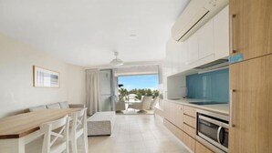 A modern kitchen with white cabinetry, a wooden countertop, and a built-in oven. There is a dining area with a white table and chairs, and a view of the outdoors through a glass door. - Salt Yeppoon (Yeppoon)