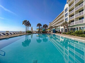 A pool with clear water, surrounded by a building with balconies and palm trees.