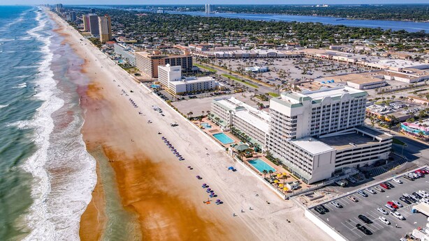 A coastal cityscape with a beachfront hotel featuring multiple floors, a swimming pool, and a parking lot. - Beachfront Studio - Sleeps 4 (Daytona Beach)
