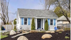 A two-story house with a front porch, a small garden with rocks and shrubs, and a blue chair in the yard. - Remodeled Jamestown beach cottage within walking distance to beach (Jamestown)