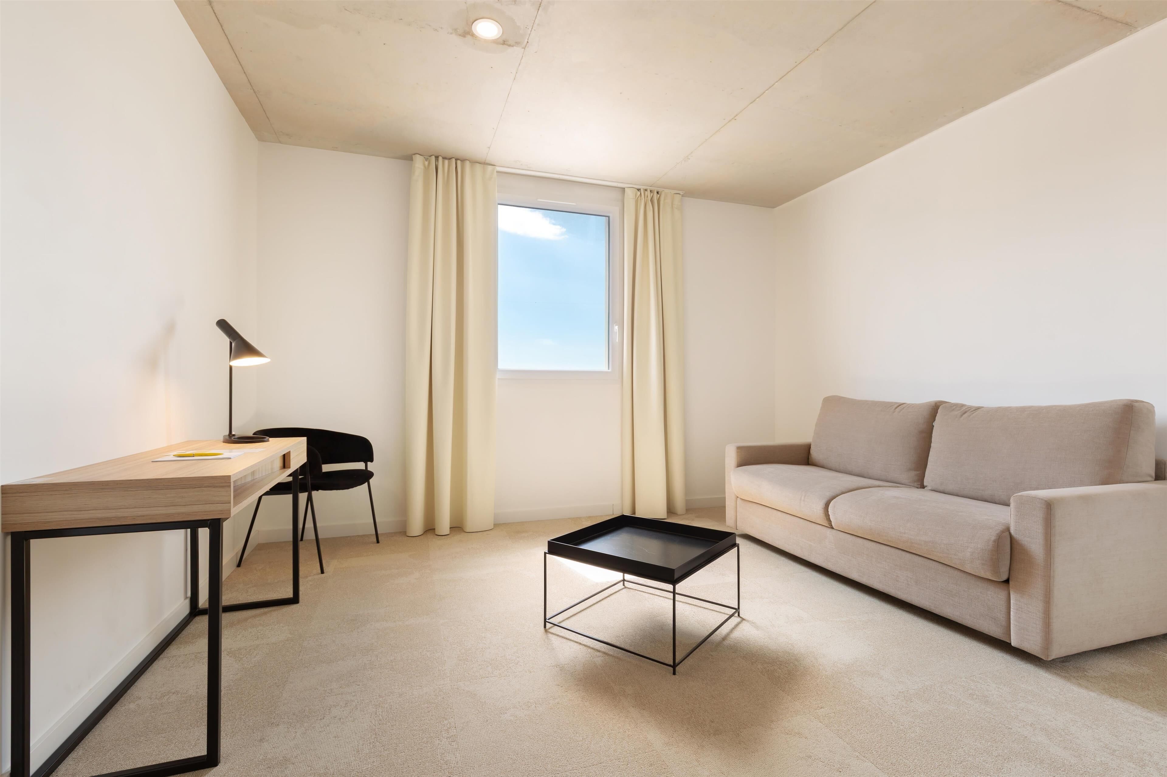A minimalist living room with a wooden desk, a black chair, a beige sofa, and a glass-top coffee table.