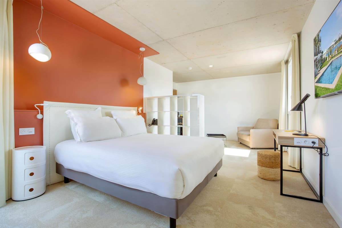 A modern hotel room with a large bed, a bedside table, a lamp, and a view of a pool.