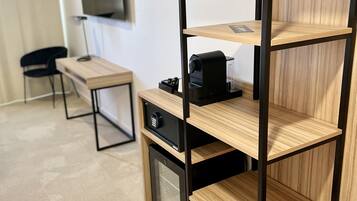Una stanza con una scrivania in legno, una sedia nera, un televisore fissato al muro e un mobiletto contenitore con una macchina da caffè.