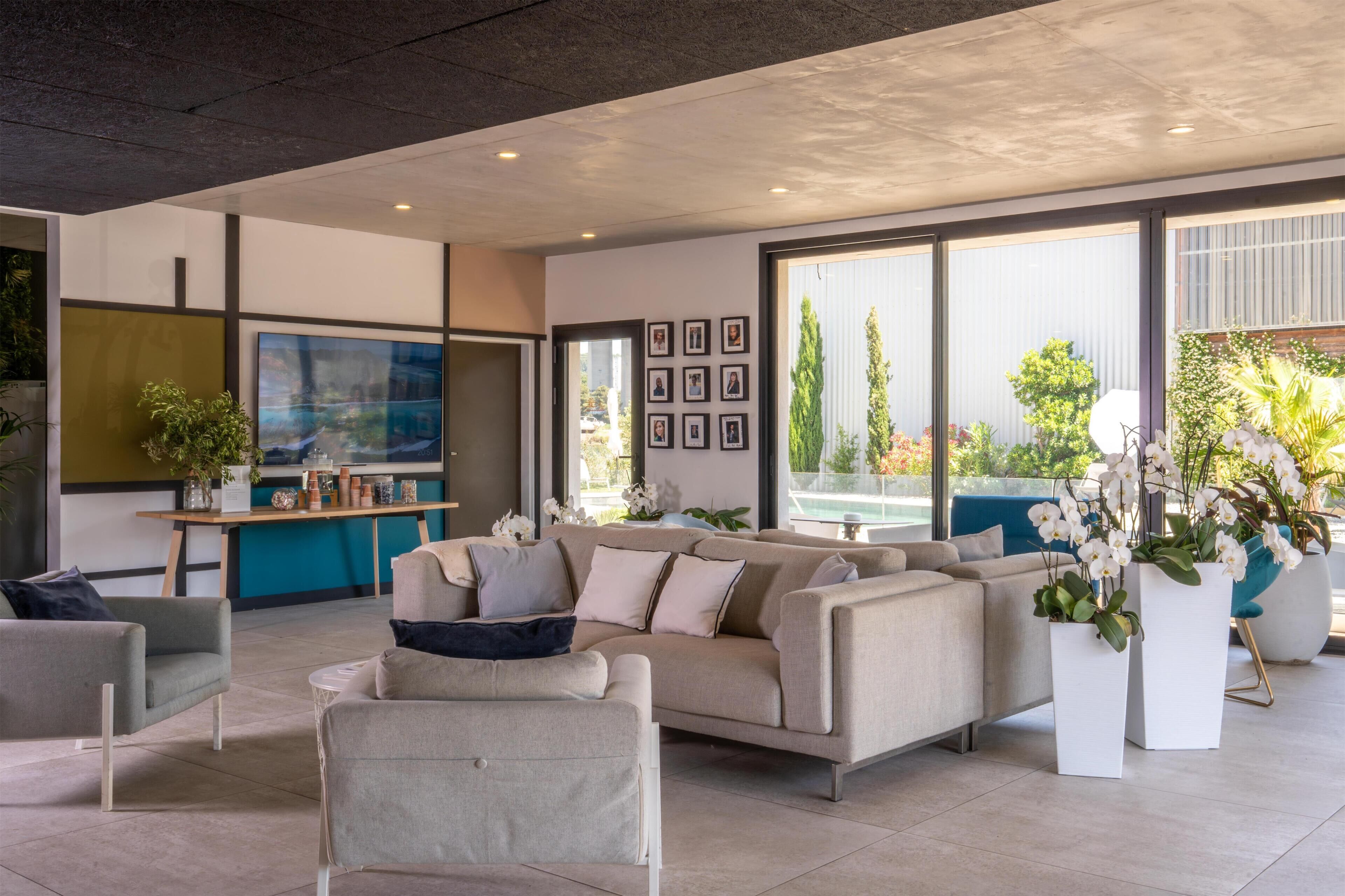 Un salon moderne comprenant un canapé, des fauteuils, une table basse et un coin repas avec une table et des chaises. De grandes fenêtres offrent une vue sur l’extérieur.