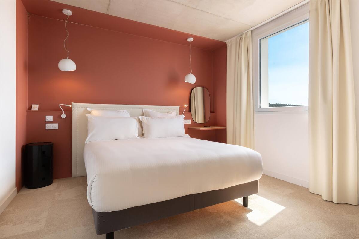 A modern bedroom with a large bed, bedside lamps, a nightstand, and a window with curtains.