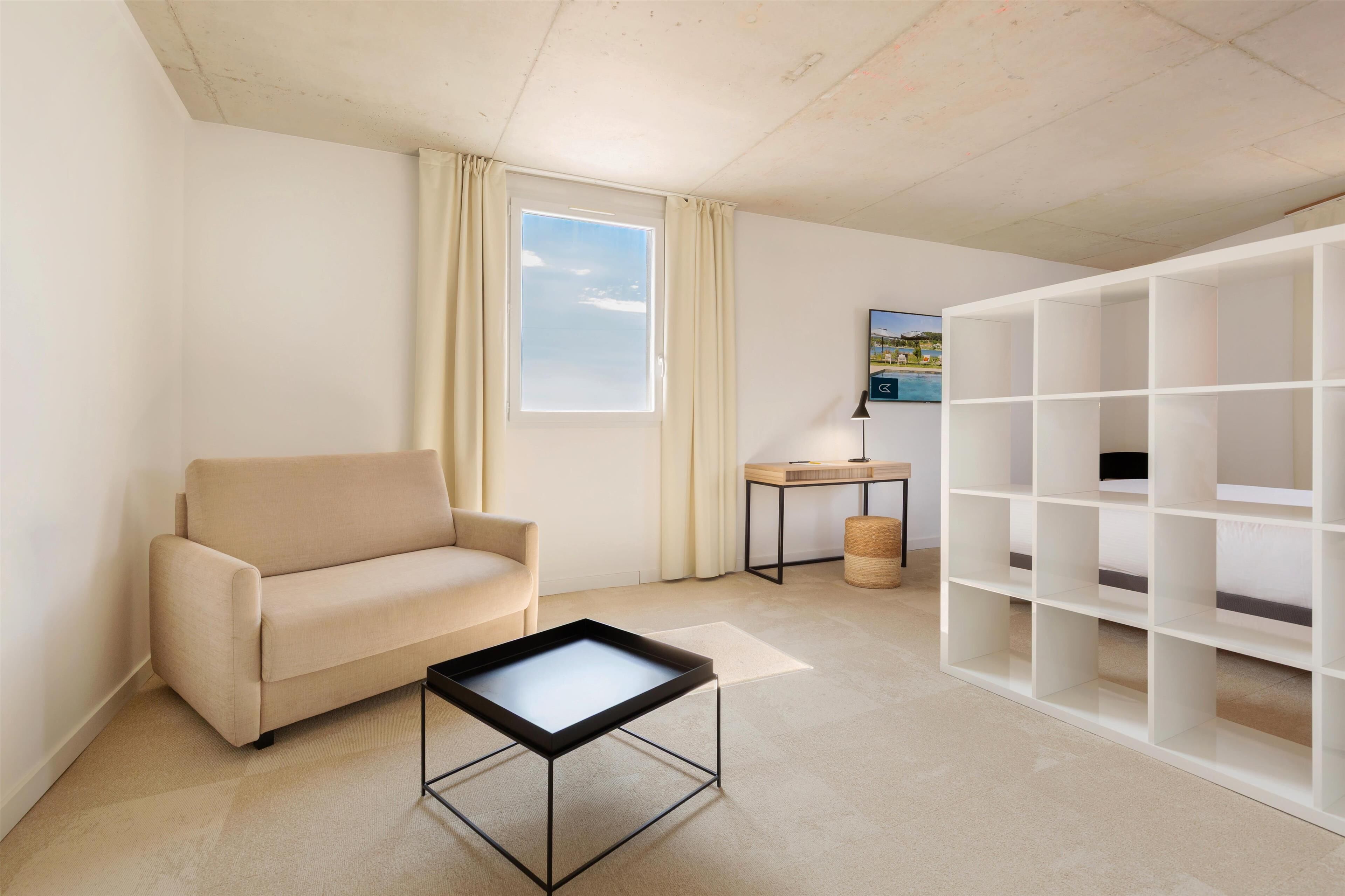 A modern living room with a beige sofa, a small coffee table, a white shelving unit, and a window with curtains.