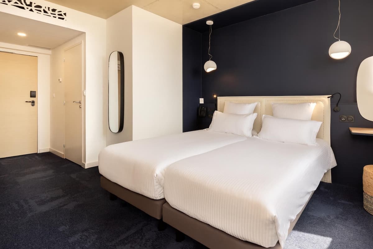A modern hotel room with a large bed, a bedside table, a mirror, and a wall-mounted light fixture.