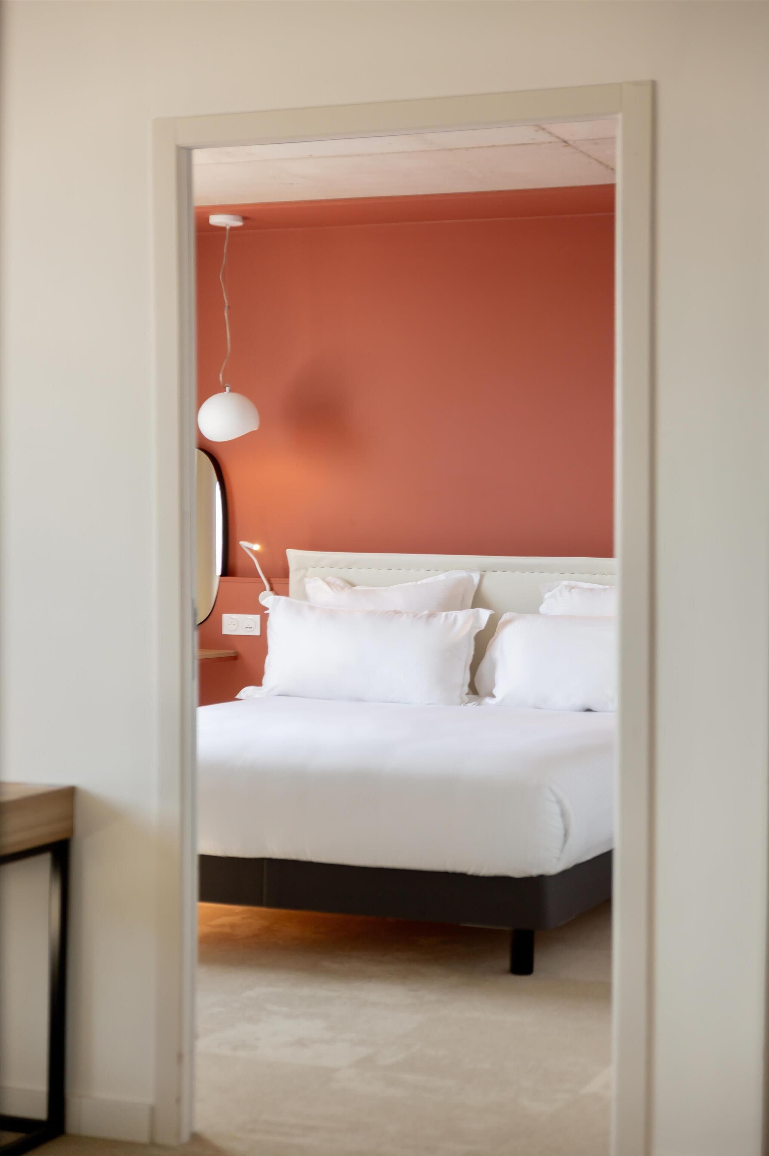 A bedroom with a bed, white bedding, a hanging light fixture, and a wall-mounted mirror.