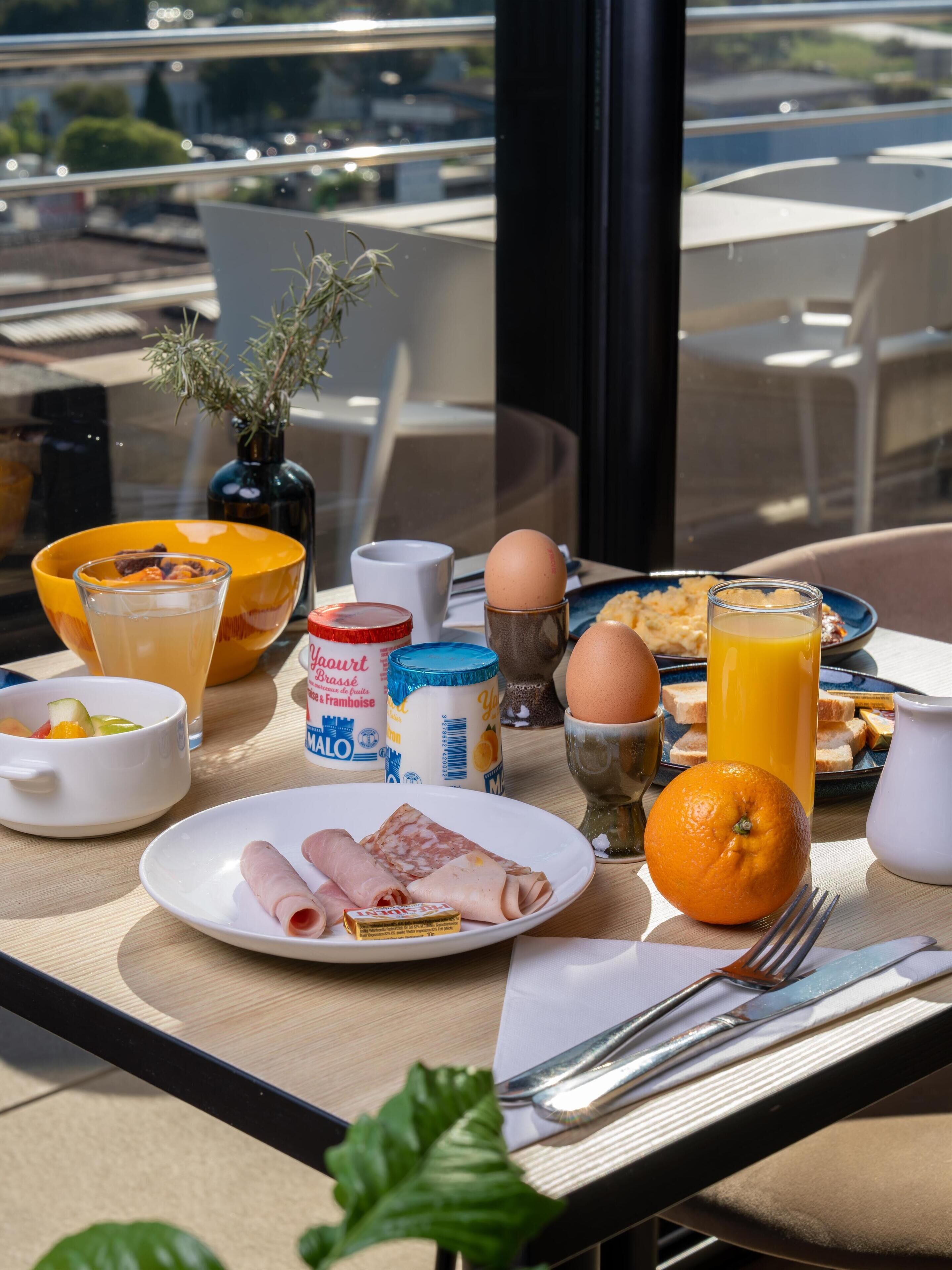 Una mesa de desayuno con alimentos, un vaso de jugo de naranja, un tazón de fruta y una vista de la ciudad a través de una ventana.