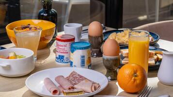 A breakfast table with food items, a glass of orange juice, a bowl of fruit, and a view of the city through a window.