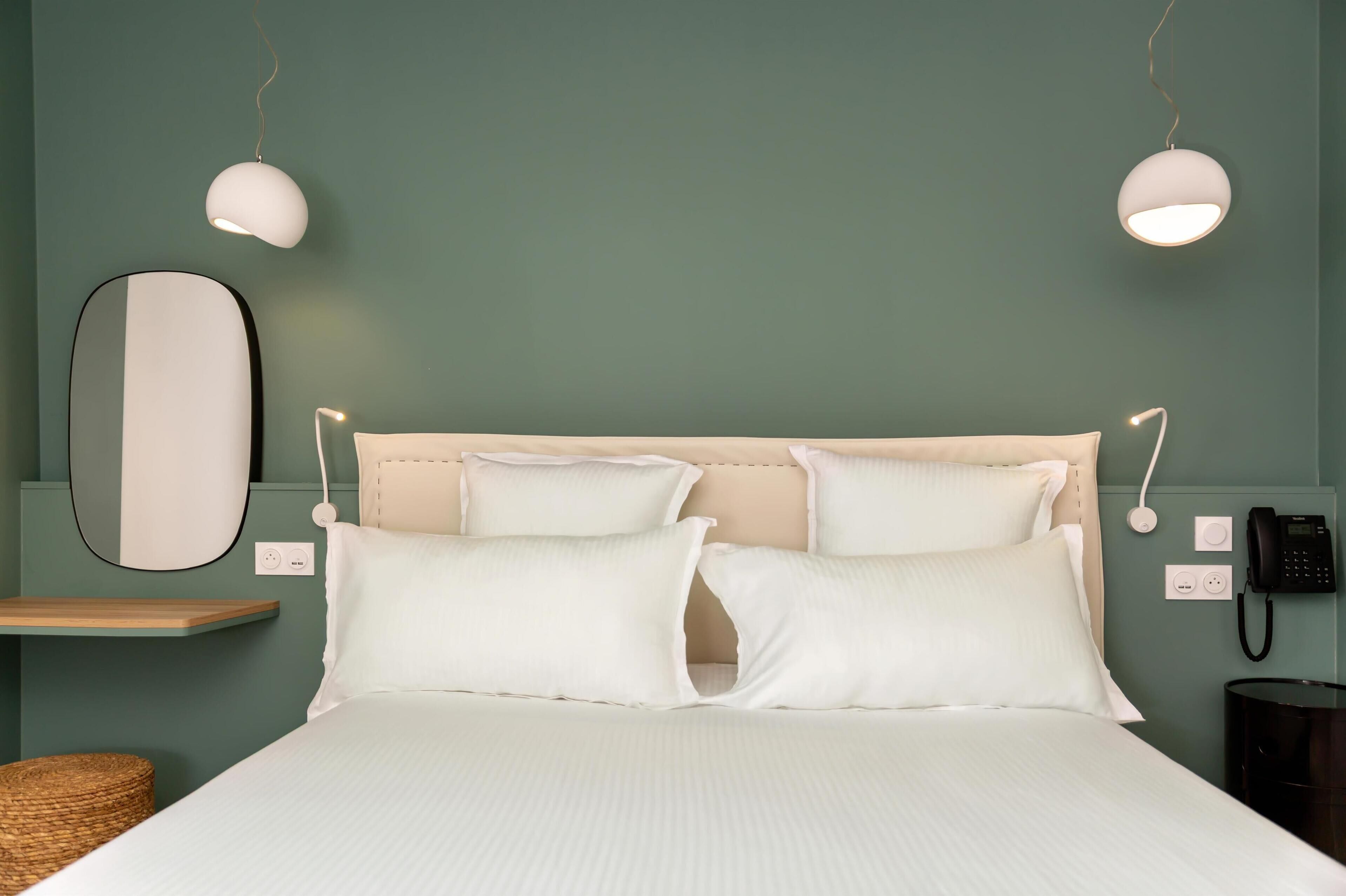 Ein ordentlich bezogenes Bett mit weißen Leinen und Kissen, ein Nachttisch mit Lampe, ein Wandtelefon und ein Spiegel.
