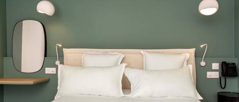 Una cama bien tendida con sábanas y almohadillas blancas, una mesita de noche con una lámpara, un teléfono en la pared y un espejo.
