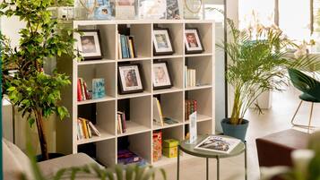 A modern living room with a white bookshelf filled with books, framed photos, and decorative items. There are potted plants and a small table with a magazine.