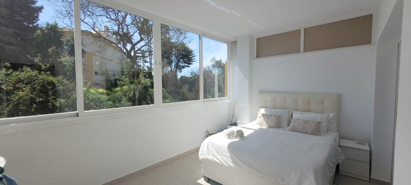 A bedroom with a large window, a bed with white bedding, and a small dog on the bed.