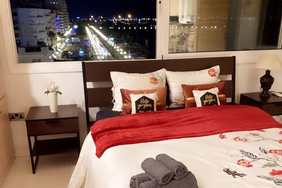 A hotel room with a large window offering a nighttime city view. The bed is neatly made with a red blanket and floral bedspread. There are two pillows with text, a small vase with flowers, and a folded towel on the bed.