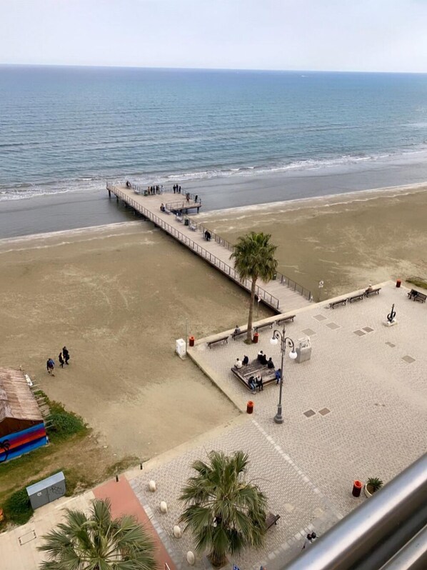 Une plage avec une longue jetée qui s’avance dans la mer, une zone sablonne agrémentée de bancs et de palmiers, et une vue dégagée sur l’océan.