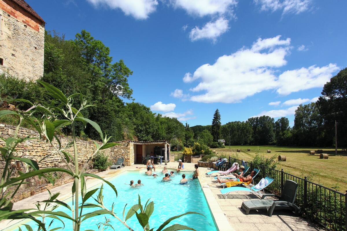 A swimming pool with people sunbathing and swimming, surrounded by greenery and a stone building.
