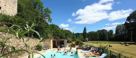 A swimming pool with people sunbathing and swimming, surrounded by greenery and a stone building.