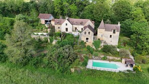 A large, multi-story stone building with a swimming pool, surrounded by dense greenery.