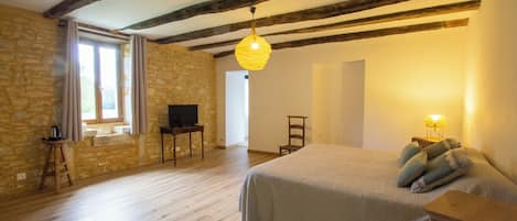 Une chambre avec un mur en pierre, des poutres apparentes, un lit avec des oreillers, une télévision, une petite table et une lampe.