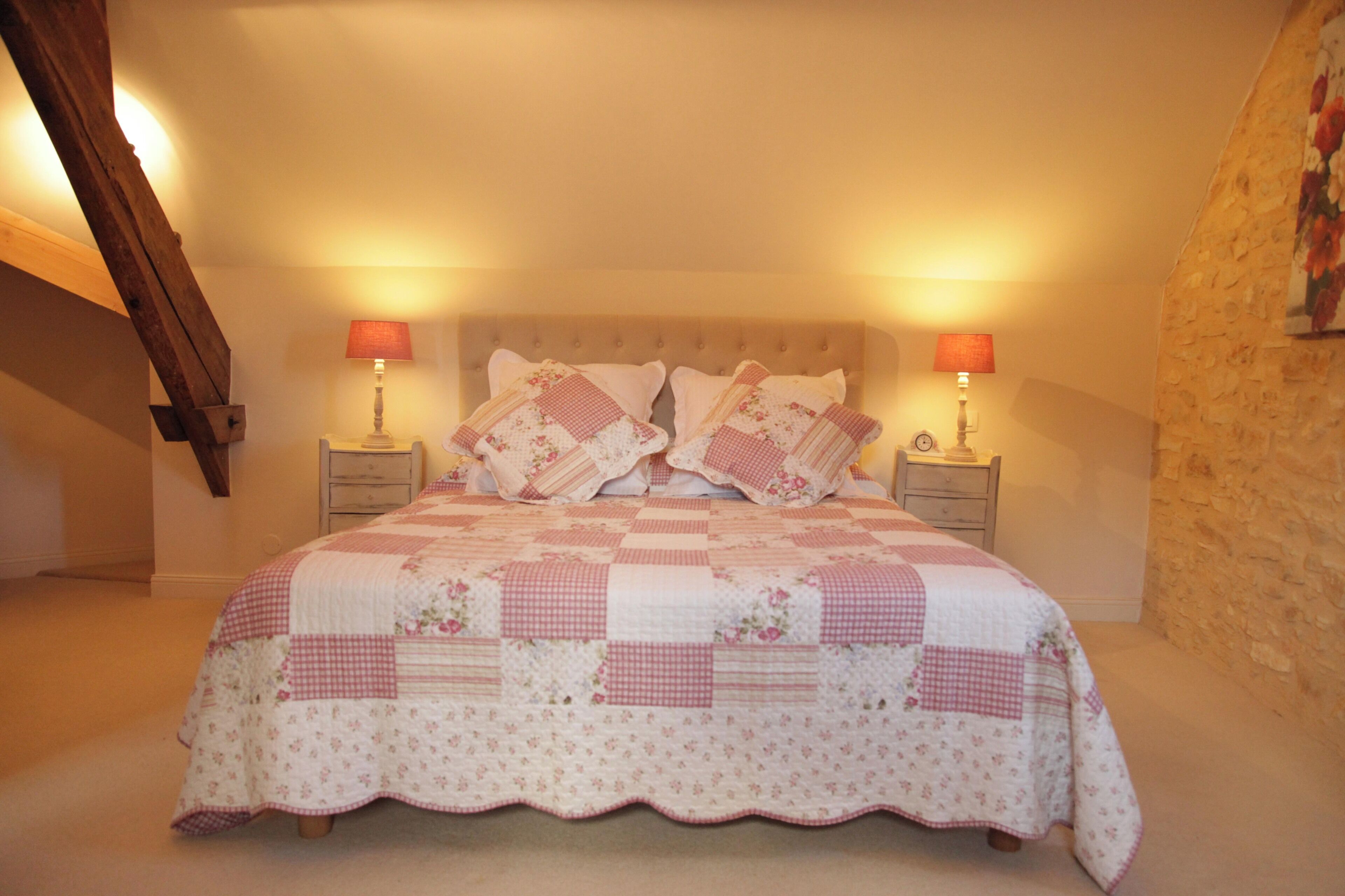 Un dormitorio con colcha acolchada, dos mesitas de noche con lámparas, viga de techo de madera y pared de piedra.