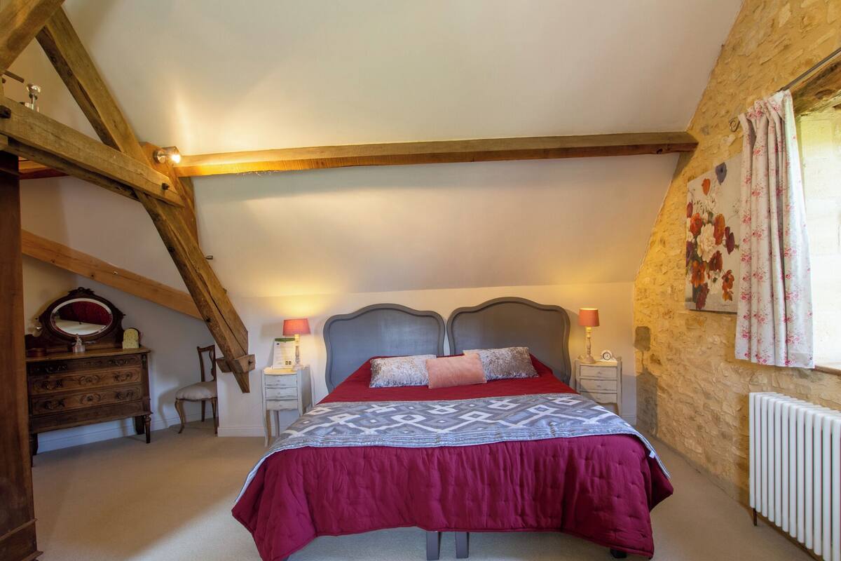 A bedroom with a high ceiling, exposed wooden beams, a bed with a patterned bedspread, bedside lamps, a dresser, and a radiator.
