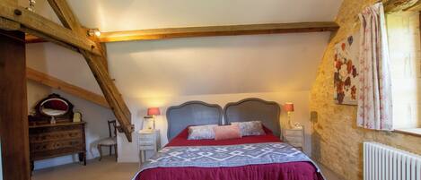 Un dormitorio con techo alto, vigas de madera a la vista, cama con colcha estampada, lámparas de noche, cómoda y radiador.